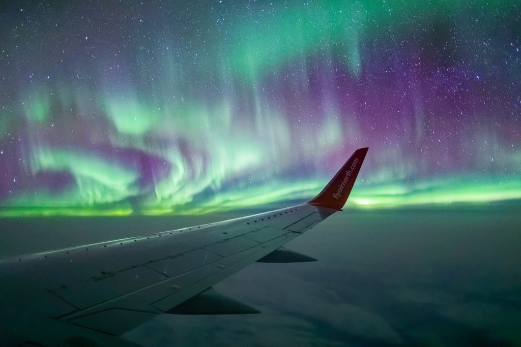 Airplane wingtip seen at night surrounded by green and purple aurora borealis
