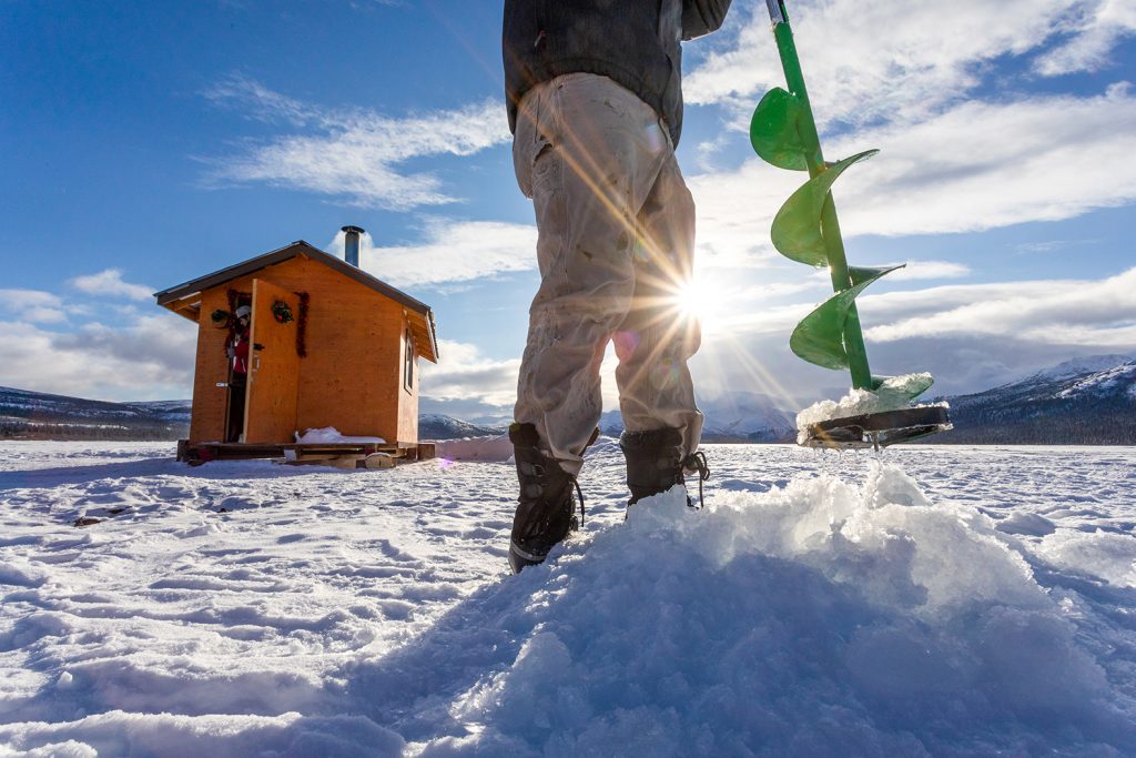 A man uses an auger to drill a hole for ice fishing as the sun sets