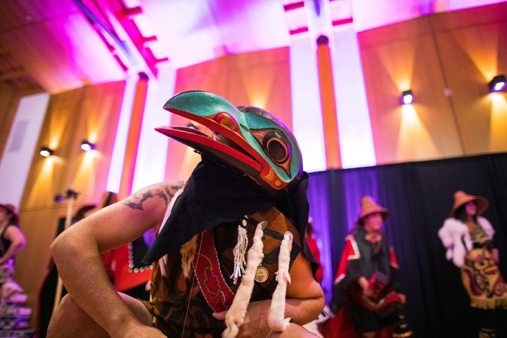 An Indigenous dancer wearing a carved mask performs surrounded by pink-tinted lights