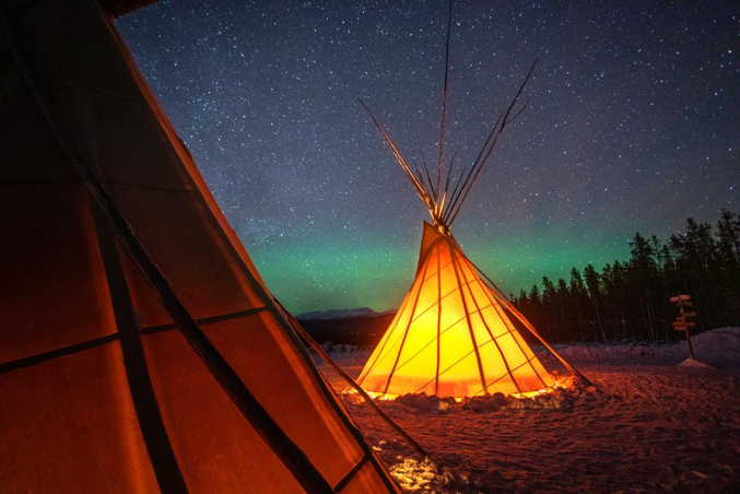A teepee, lit from within, stands out against a starry night sky with aurora borealis visible on the horizon