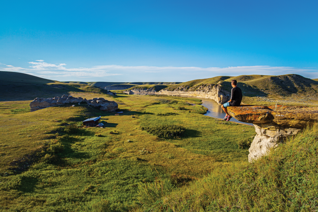 A man sits on a rocky ledge overlooking a river and expansive grasslands.