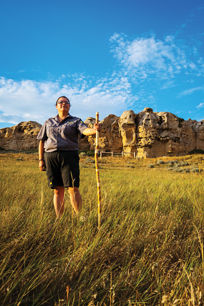 A woman stands in grasslands holding a wooden staff, cliffs in the background