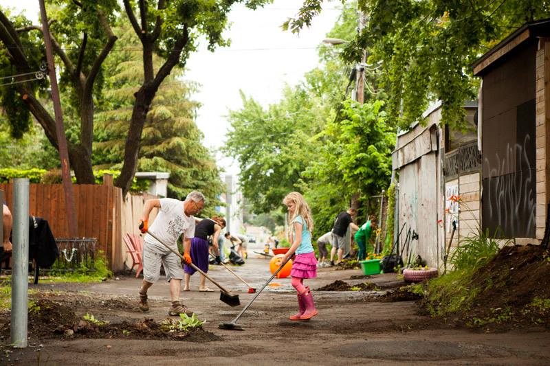 A man and a girl sweep the ground of an alley, other people work in the background